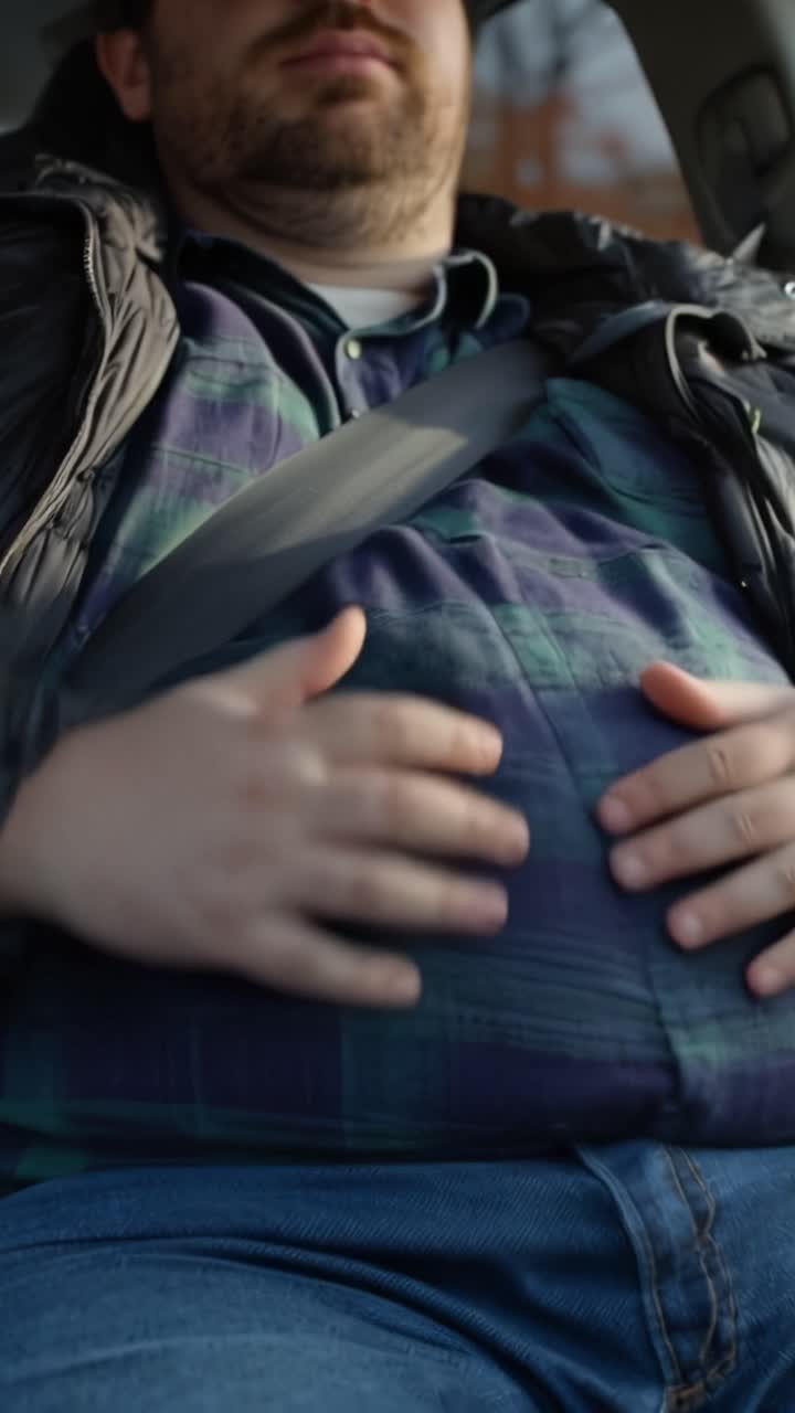 Hombre duerme en el coche, con el cinturón de seguridad cruzado sobre el abdomen. Su mano descansa sobre su estómago. Parece cansado, inconsciente de su entorno. Este video sugiere descanso, relajación o fatiga. Uso en viajes, bienestar.