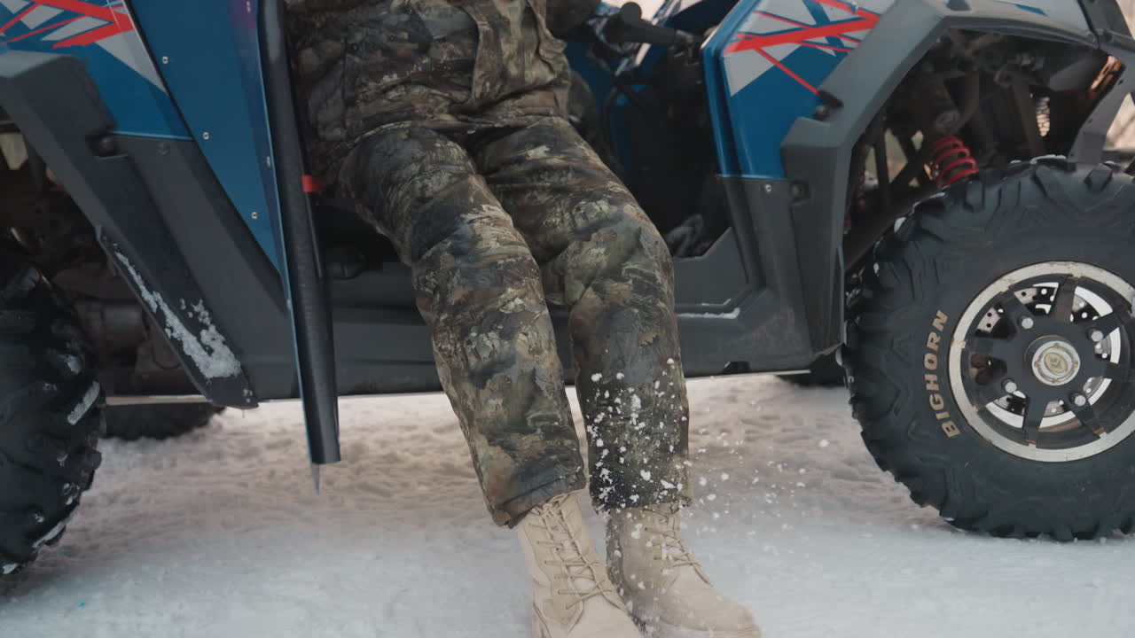 Pilota in mimetica si prepara sul suo ATV, fissando casco e guanti all'interno di un veicolo utilitario blu sul ciglio di una strada innevata al crepuscolo. Un'espressione concentrata, pneumatici robusti che sollevano la neve, creando un'atmosfera avventurosa e tesa.