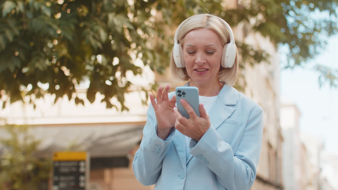 feliz y despreocupada mujer de negocios madura con teléfono inteligente escuchando música en auriculares en la calle de la ciudad