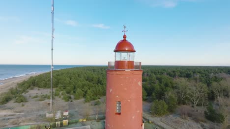 aerial establishing view of red colored akmenrags lighthouse, baltic sea coastline, latvia, white sand beach, calm sea, sunny day with clouds, medium ascending drone orbit shot