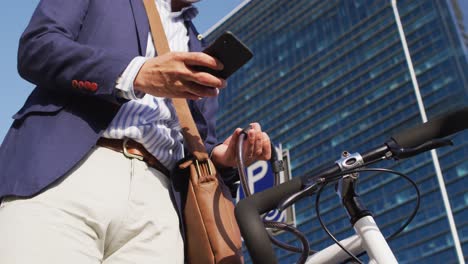 Hombre-Asiático-Con-Mascarilla-Usando-Un-Teléfono-Inteligente-Mientras-Bloquea-Su-Bicicleta-En-El-Parque-Corporativo