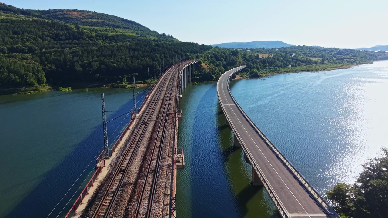 Udsigt over en flod og jernbanespor med en motorvej langs vandet nær grønne bakker på en solrig dag