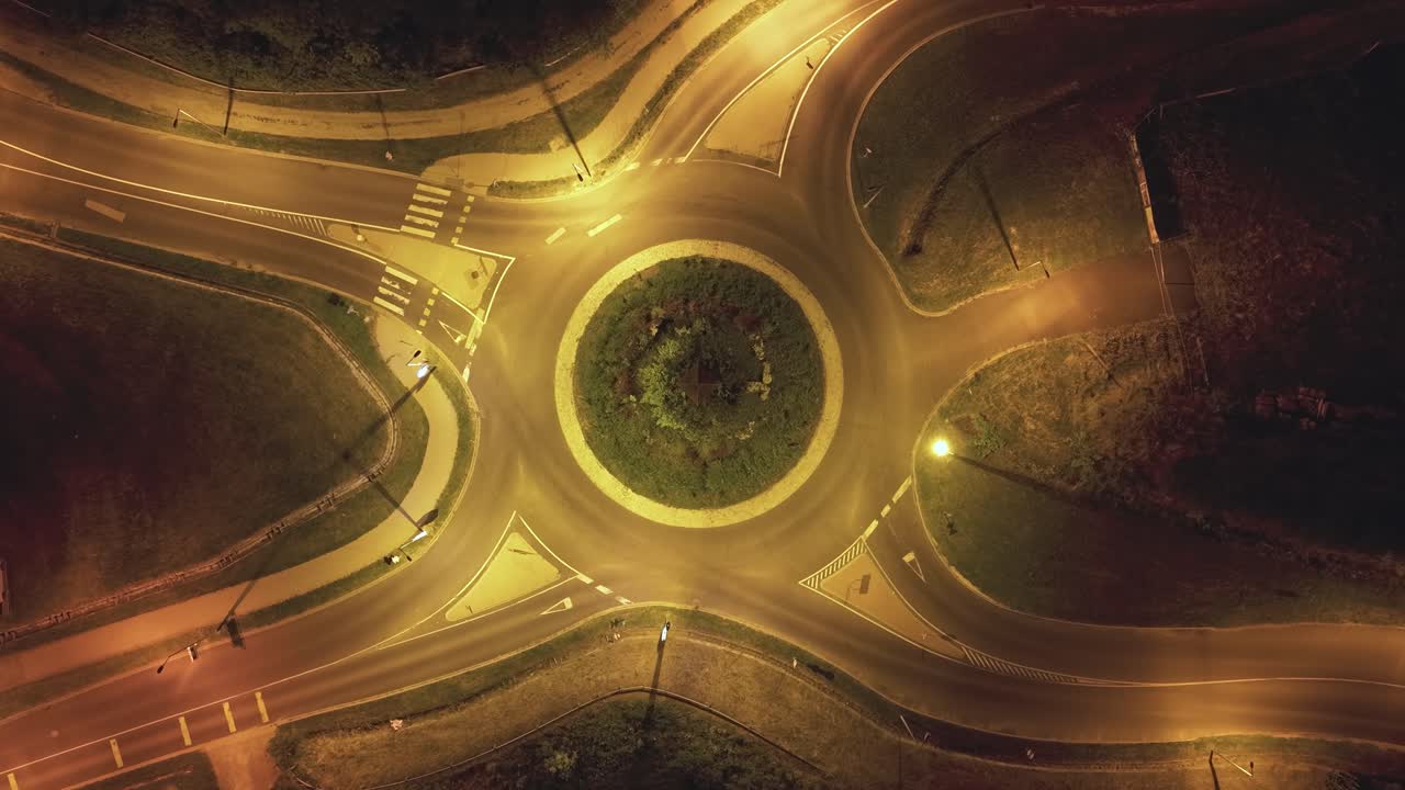 Aerial view of a roundabout intersection illuminated by streetlights at night. Time lapse of moving vehicles create dynamic lines, emphasizing the structure's symmetry and the urban traffic flow
