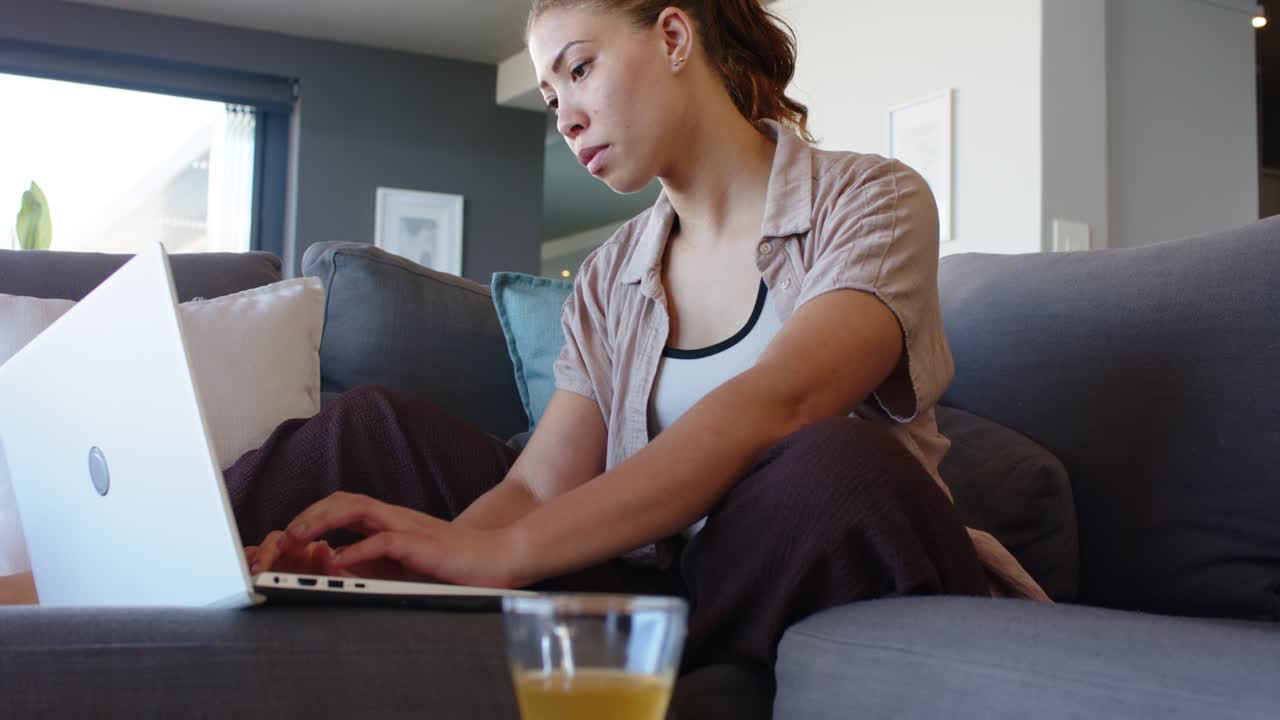 Afroamerikansk kvinde sidder på sofaen, justerer sin bærbare computer for at taste, med en orange drik i nærheden.