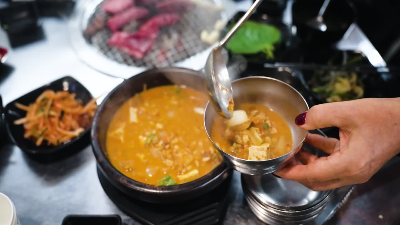 Close-up shot of a steaming earthenware pot of Doenjang Jjigae Korean stew being scooped with a ladle into a silver metal bowl next to side dishes