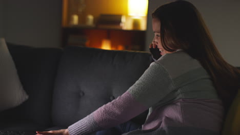 mujer sonriente sentada en el sofá en casa por la noche hablando en el teléfono móvil y viendo una película o un programa en la computadora portátil 4