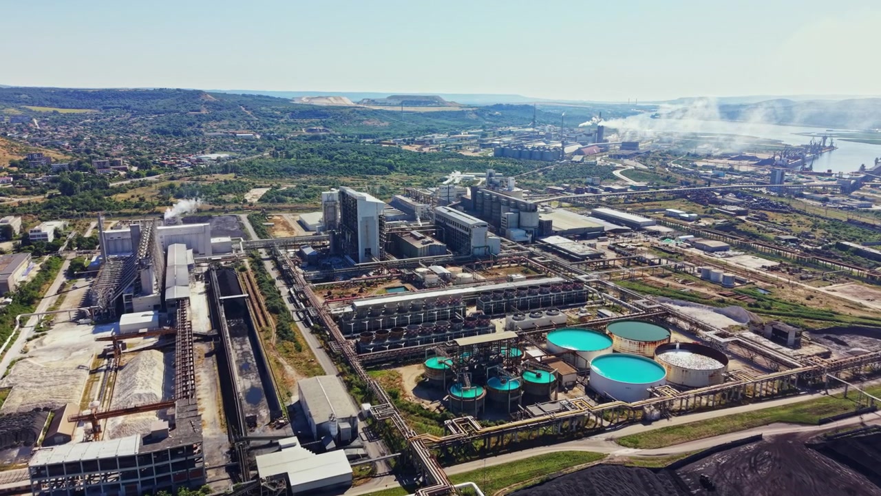 Sito industriale in un'area rurale con grandi edifici, serbatoi e macchinari, circondato da colline e un fiume sullo sfondo, in pieno giorno.