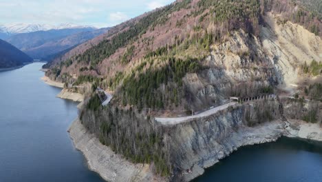 winding road along a scenic mountain lake surrounded by forested hills, aerial view