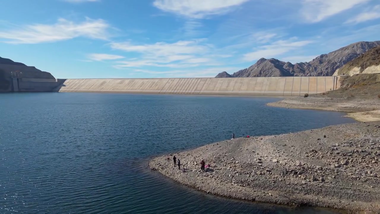 Visitors stand along the rocky shoreline of Punta Negra Reservoir below the concrete-faced rockfill Punta Negra Dam on the Zonda River, Ullum, San Juan, Argentina, fast drone forward flyover.
