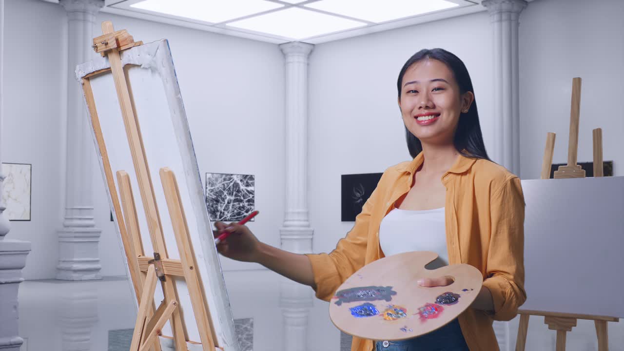 아시아 여성 예술가 가 갤러리에서 오일 페인트와 브러쉬로 그림을 그리는 동안 캔버스를 가리키고 있습니다.