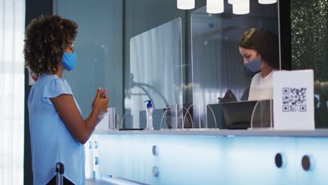 Mixed-race-woman-and-female-receptionist-wearing-face-masks-at-hotel-reception-desk