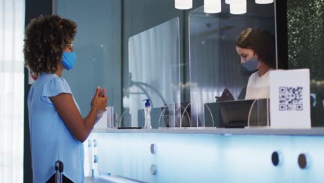 mujer de raza mixta y recepcionista llevando máscaras faciales en el mostrador de recepción del hotel