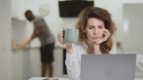 mujer blanca sorprendida leyendo noticias en una computadora portátil en una cocina abierta.