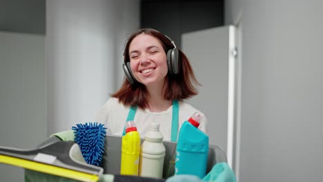 a cheerful brunette cleaning lady listens to music in black wireless headphones walks along the apartment corridor and carries a gray plastic basin with cleaning tools and detergents