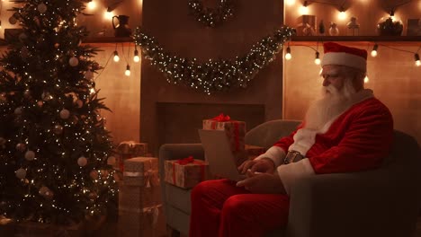 santa claus is sitting on a comfortable soft couch, holding a laptop in his lap and typing on it.