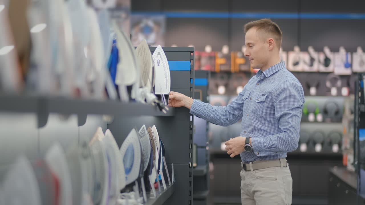 A young handsome man chooses an iron in a consumer electronics store for his new home. Studies the characteristics. Takes samples from the counter and examines the quality of the devices