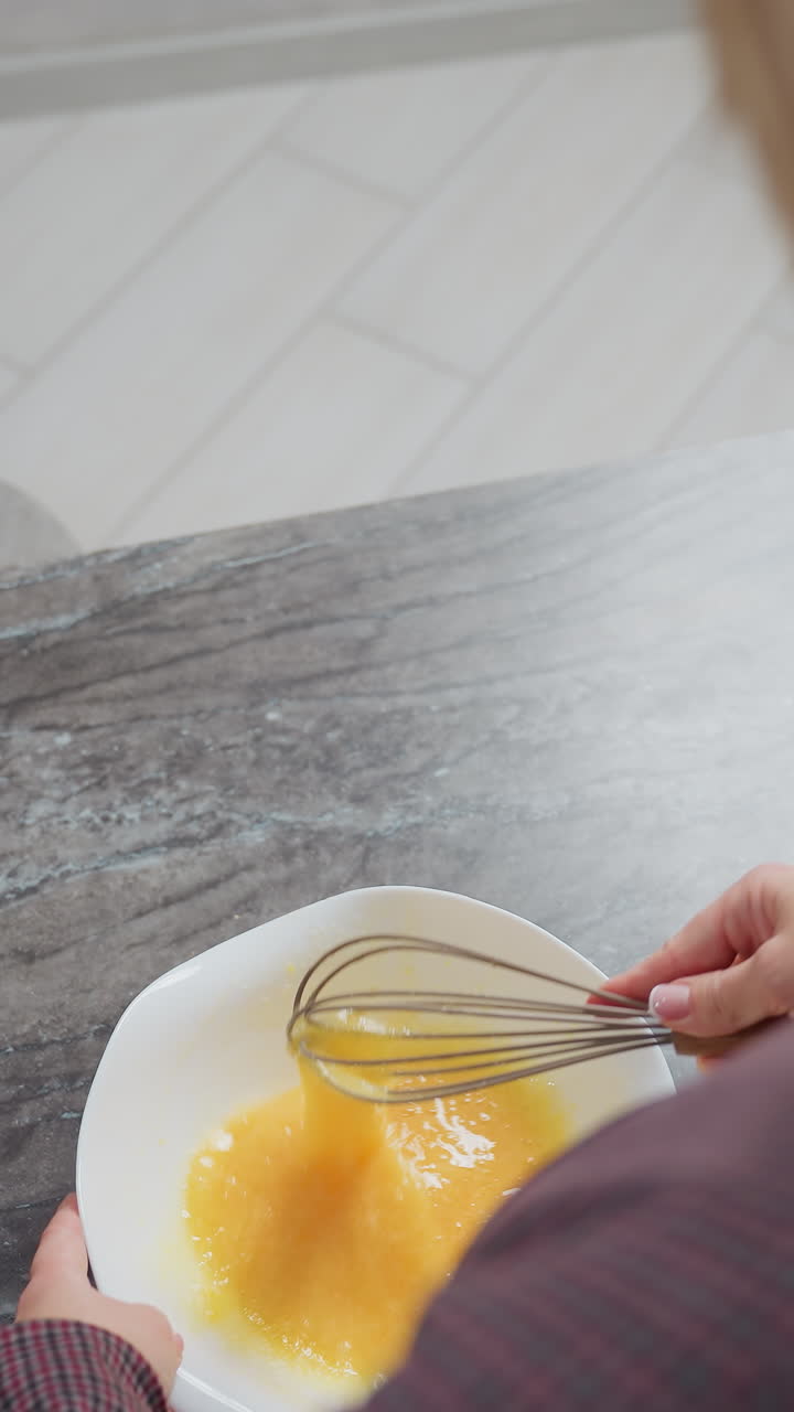 vista trasera de la dama con el cabello dorado usando un vestido a cuadros agitando el huevo en el cuenco, ella levanta el batidor ligeramente antes de continuar mezclando, con una encimera lisa y iluminación suave