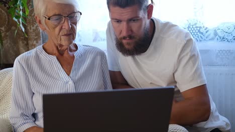 happy senior mother, adult son sitting in living room at sofa and using laptop trying to make online payment. retirement lifestyle, family spending quality time at home. middle-aged man helping mum