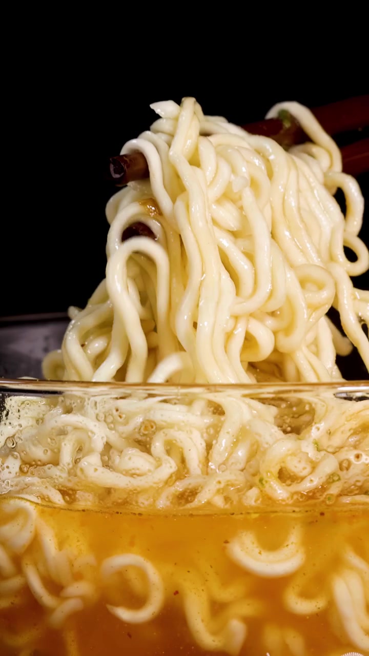 Le bacchette mescolano noodles istantanei fumanti in una ciotola di vetro trasparente piena di brodo, sotto un'illuminazione drammatica da studio e su uno sfondo nero solido.