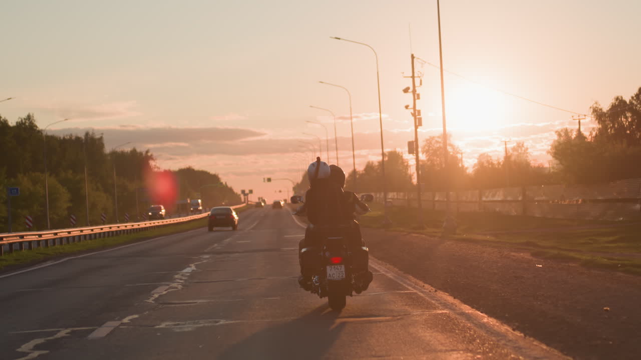 Back view of motorcycle rider with passenger traveling on open highway during golden sunset, bright sunlight casting warm glow across road and vehicles