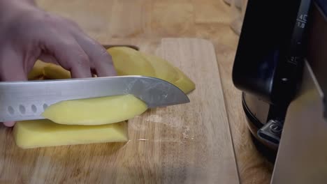 Toma-De-Control-Deslizante-En-Cámara-Lenta-De-Cortar-Una-Papa-En-Papas-Fritas-En-Una-Tabla-De-Cortar-De-Madera-En-La-Cocina