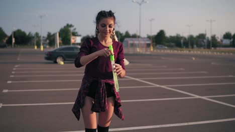 Young-Playful-Hipster-Girl-With-Long-Braids-Blowing-Bubbles-On-The-Parking-By-The-City-Mall-During-Sunset