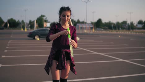 young playful hipster girl with long braids blowing bubbles on the parking by the city mall during sunset