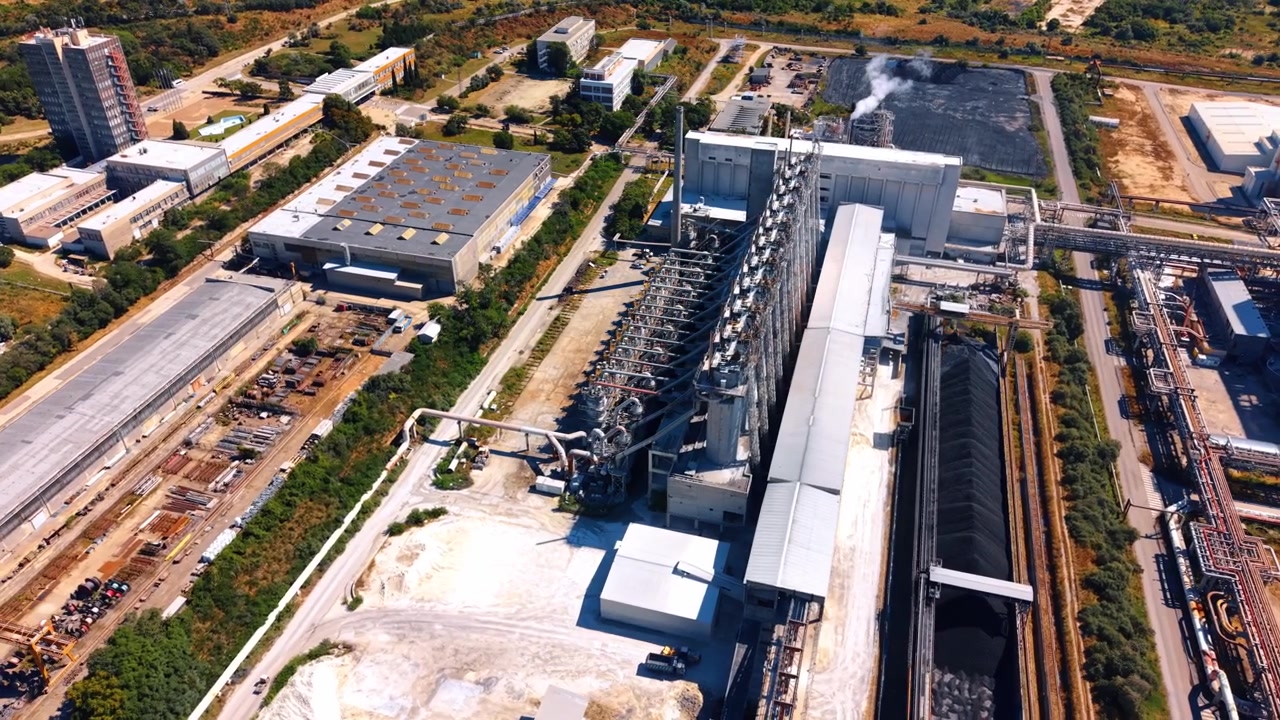 High angle aerial view of a large industrial plant complex. Railway tracks and coal storage piles lay adjacent to steel processing buildings on sunny day.