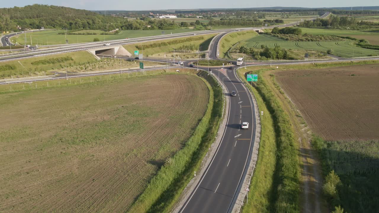 Aerial video of a highway roundabout in rural Hungary, with visible traffic including cars, a motorcycle, and a bus. The roundabout is surrounded by green fields and cultivated farmland.