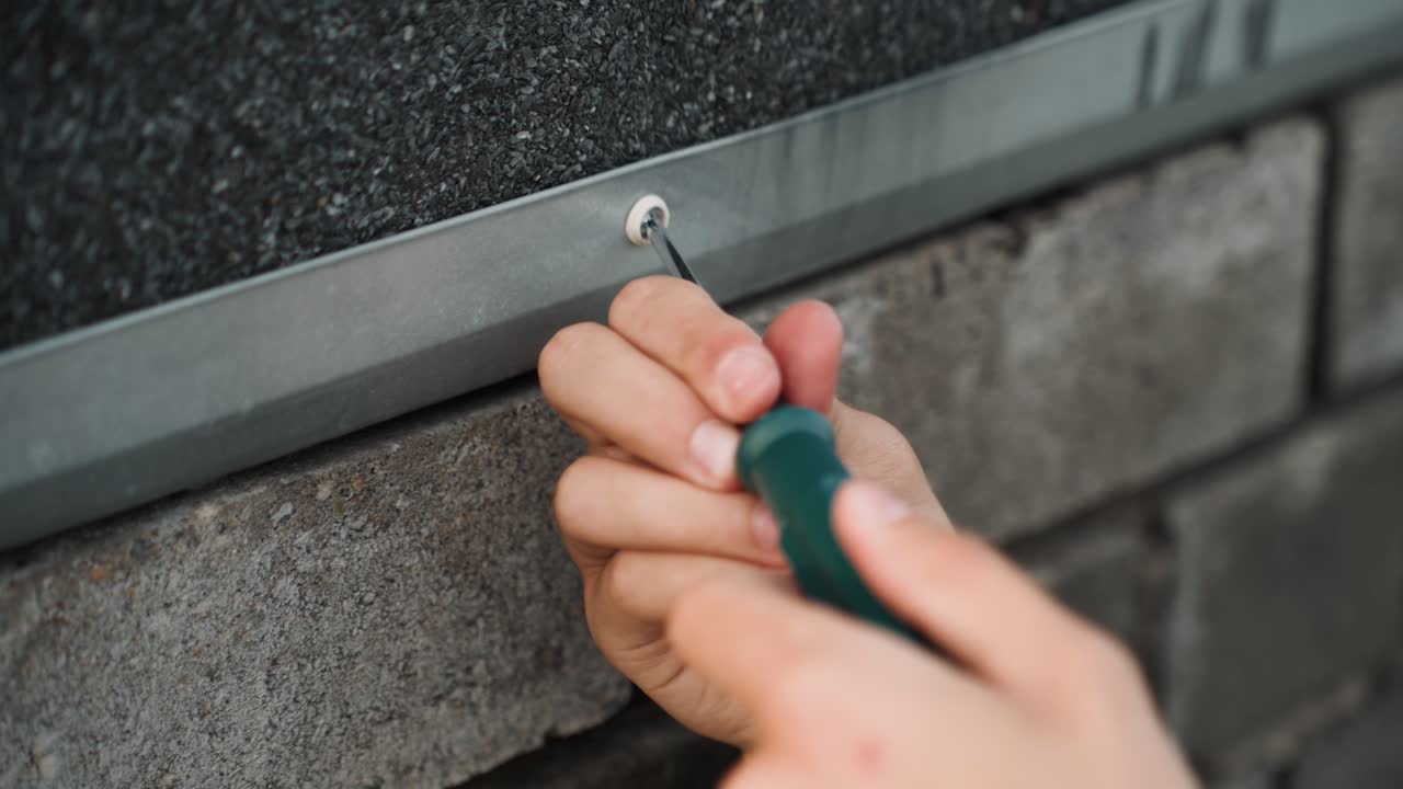 Detail shot shows hands tightening screw on metal wall, professional technician performing exterior maintenance on brick facade, torque applied with green handled screwdriver, close attention