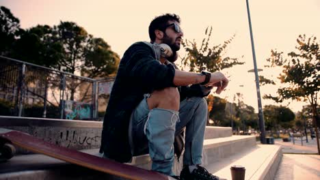 young skateboarder with headphones and coffee relaxing at skateboard park