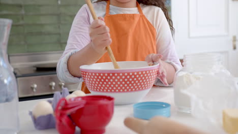 Happy-biracial-girl-with-long,-curly-hair-mixing-dough-in-bowl-and-smiling-in-sunny-kitchen