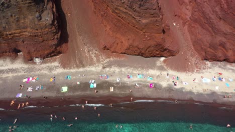 Aéreo-De-Arriba-Hacia-Abajo-Volando-Lateralmente-A-Través-De-La-Playa-Roja-Con-Agua-Turquesa,-Turistas,-Montaña-Y-Arena-De-Color-Rojo-En-Santorini,-Grecia