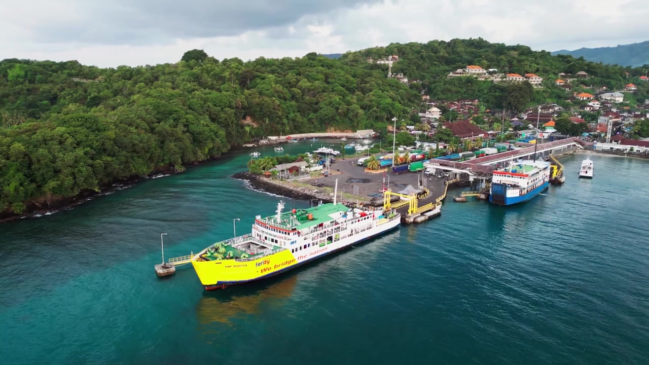 Area di carico del terminal portuale a Bali con un traghetto passeggeri ormeggiato accanto a una collina tropicale verde e acqua calma dell'oceano blu, riprese aeree con drone.