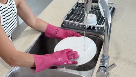 individual wearing pink gloves scrubs and rinses a white plate under running water in a modern kitchen with bright, even lighting and steady camera angle
