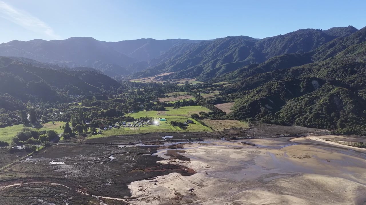 Vista aerea di una valle fluviale intrecciata e di un paesaggio montano sull'Isola del Sud della Nuova Zelanda con canali d'acqua tortuosi e campagna verde sotto un cielo azzurro limpido.