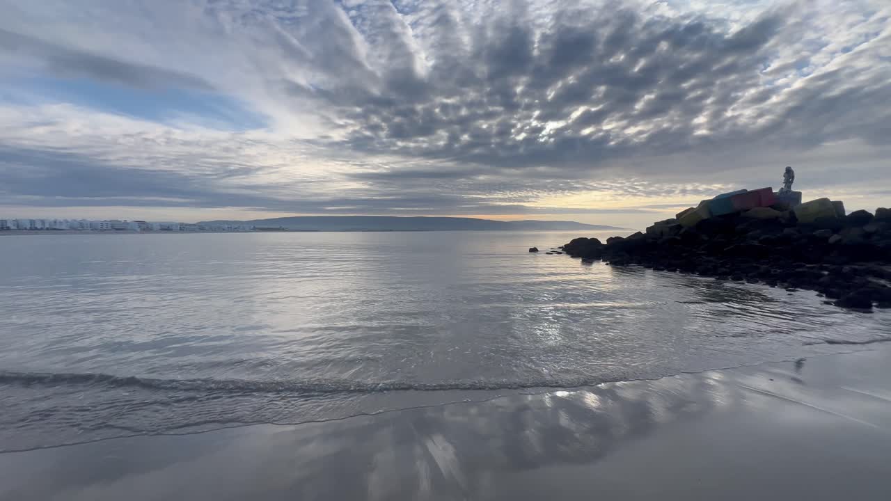 Capturing the serene beauty of a peaceful sunrise illuminating the calm waters of Barbate's beach in Spain, with gentle waves lapping against the sandy shore under a cloudy sky