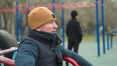 close-up of a coach sitting with hands resting on workout equipment, looking tired, as he shakes his head, in the blurred background, a boy stands near blue poles