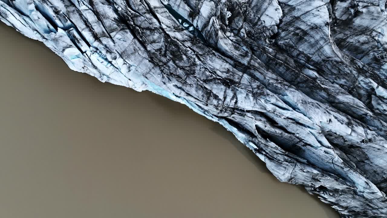 Close-up aerial view of Vatnajökull glacier edge, dark ash streaks and blue crevasses meeting a muddy lagoon—dramatic Iceland meltwater and climate change concept