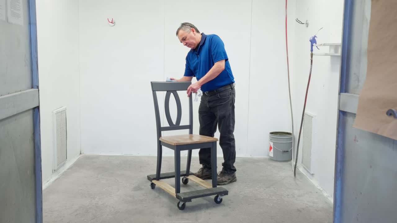 Camera locked as man sands wooden backrest of dining chair inside paint booth