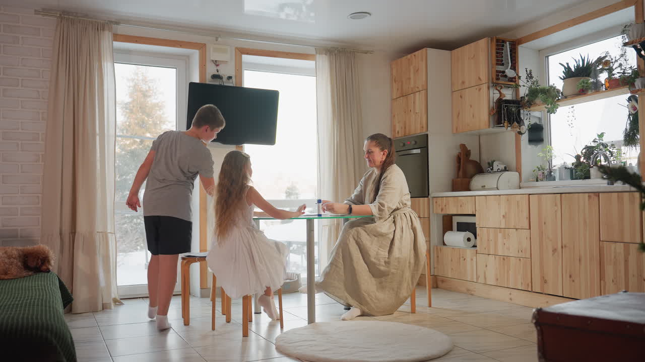 Children sitting in front of their mother, all wearing socks. The mother hands them paper while sitting at a table, background features cozy furniture and television, creating a family bonding moment