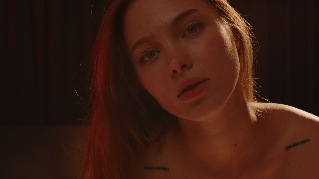 Smukt nærbillede af en attraktiv og ung kvinde, der sensuelt ser ind i kameraet. Pigen har dybblå øjne og tatoveringer på skuldrene, og hun bevæger sig baglæns i slow motion.