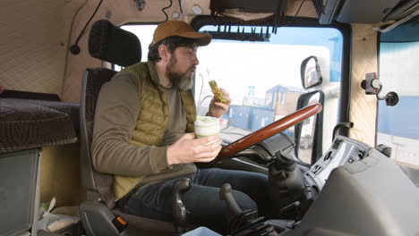 side view of worker sitting in a truck in a logistics park while eating a sandwitch and holding a drink