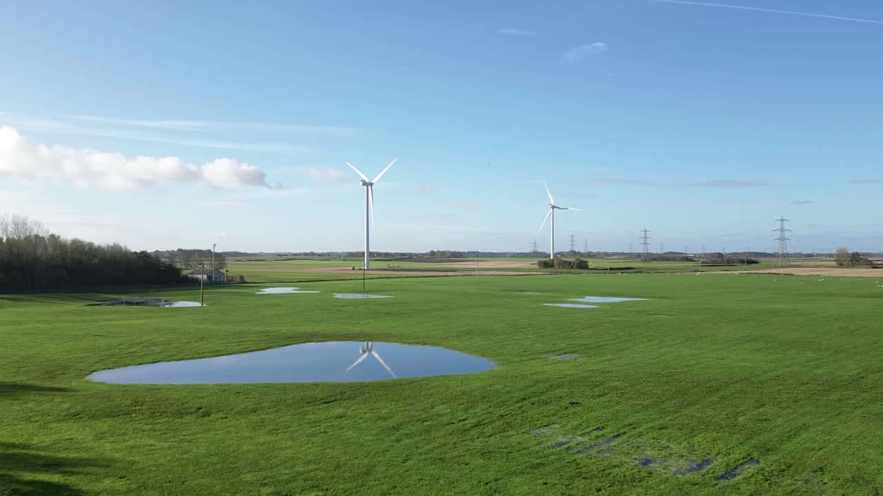 Two wind turbines rotating in a green field with one turbine’s reflection visible in a large puddle, against a near clear blue sky. Captures the harmony of wind energy and nature.