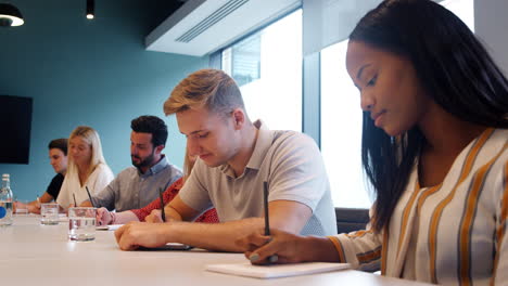 group of young candidates sitting at boardroom table making notes at business graduate recruitment assessment day