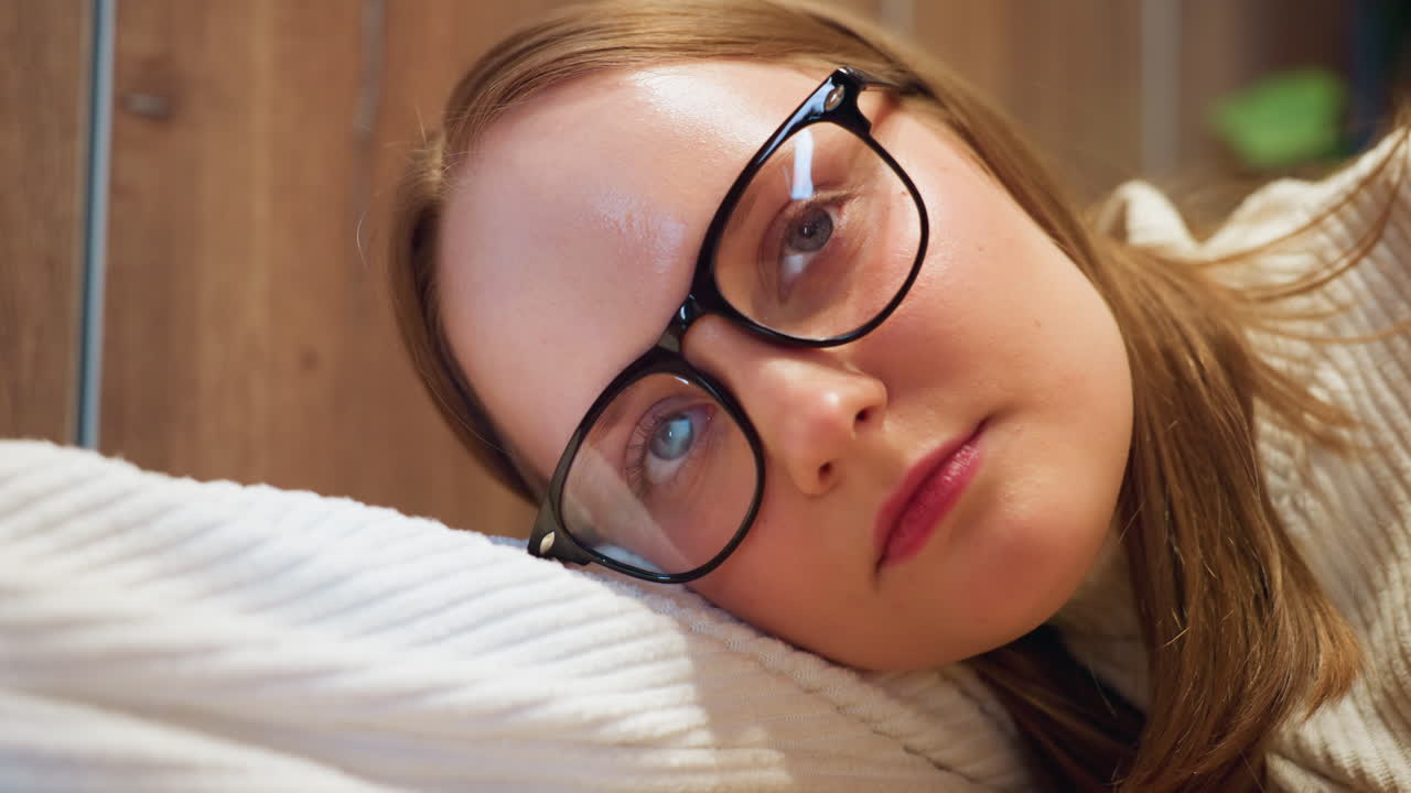 close up of woman resting head on wooden table tapping tabletop lightly with finger, wearing glasses and white sweater, showing fatigue and contemplation in indoor setting with greenery background