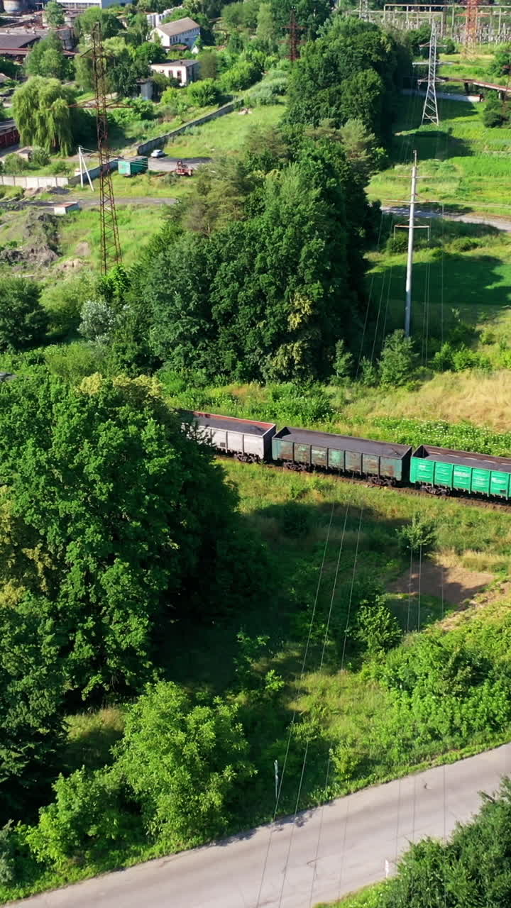 Freight train passing on railway. Cargo train with freight wagons moving among green nature in summer. Aerial view. Vertical video