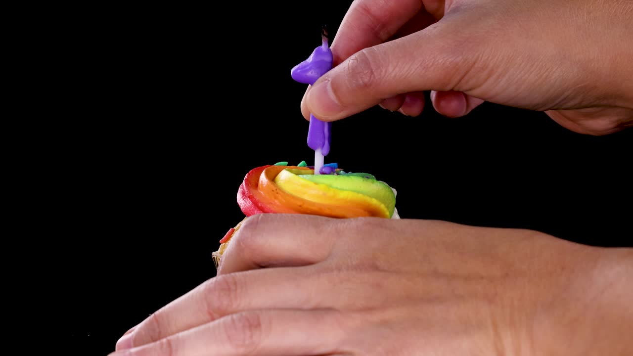 A hand inserts a purple number one candle into a rainbow-frosted cupcake against a black background, with steady lighting and close-up framing