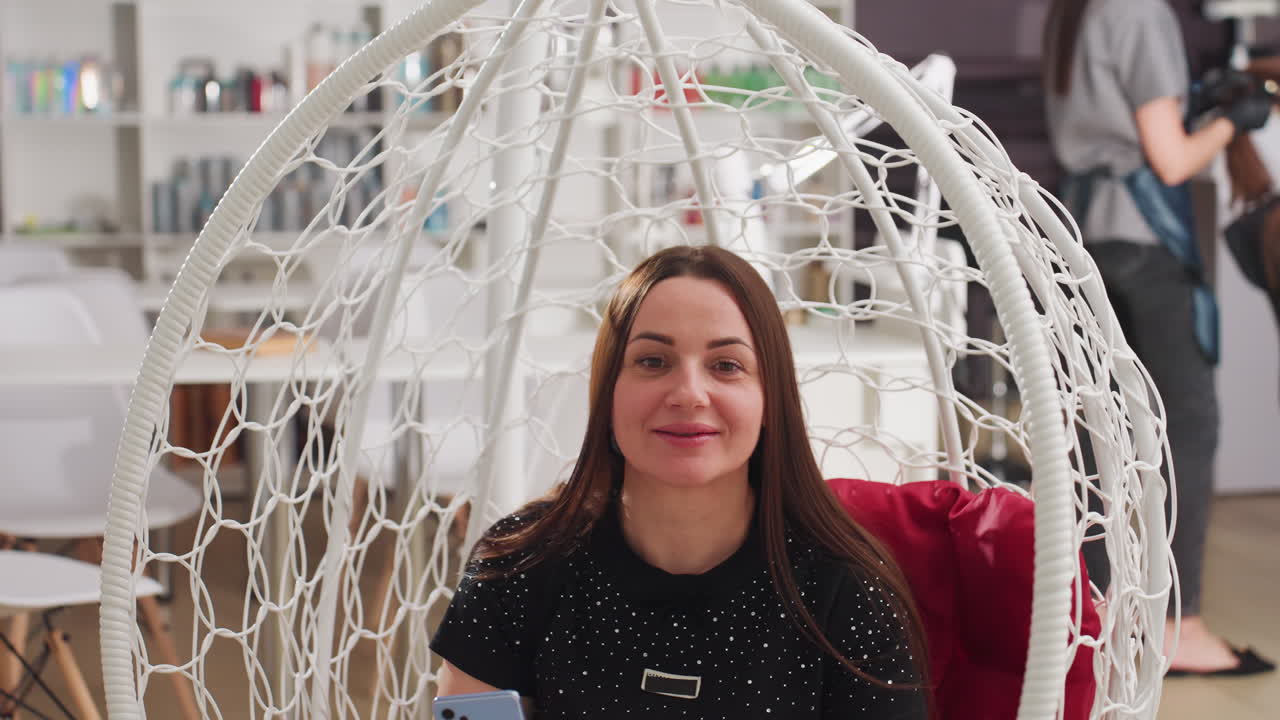 Una mujer sonriente se hace un selfi en un sillón colgante en un salón; una clienta relajada captura momentos espontáneos mientras el estilista trabaja al fondo, interior luminoso con estanterías de productos y un cómodo asiento de columpio tejido