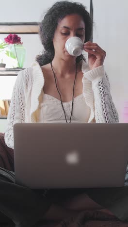 young african american woman sits with laptop on sofa and drinking coffee